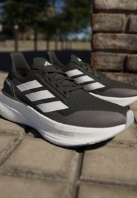 Chaussures de sport noires et grises avec trois rayures blanches, dessus en maille tricotée, semelle rembourrée et détails texturés. Conçues pour le confort et le soutien.