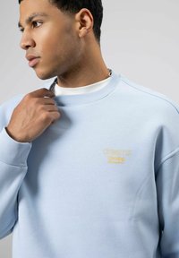 Sweatshirt bleu clair en tissu doux avec poignets et col côtelés. Présente un petit logo jaune brodé. Gros plan sur l'encolure et l'épaule.