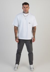 Camisa de algodón blanca con mangas cortas y logo bordado en el pecho, combinada con pantalones cargo grises con bolsillos con botones. Zapatillas blancas.