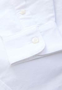 Chemise blanche en coton avec poignets boutonnés et boutons imitation perle sur une surface de tissu lisse.