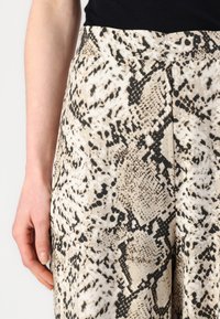 Pantalons en imprimé serpent beige et noir avec une texture douce, présentant une taille haute et un design de jambes évasées.