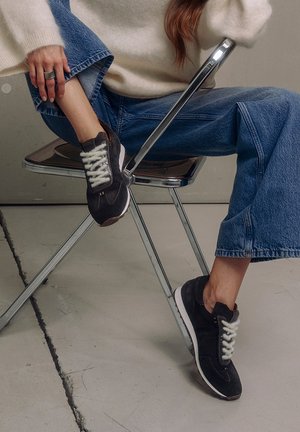 Zwarte suède sneakers met witte veters, een witte zool en een strak ontwerp. Gemaakt in combinatie met blauwe wijde jeans, terwijl ze rusten op een chroomstoel.