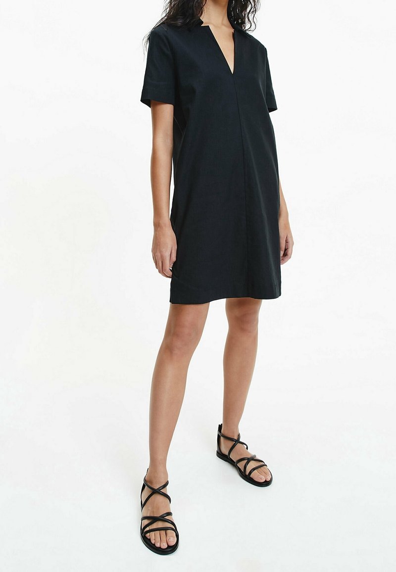 Robe noire manches courtes à col en V, avec une coupe décontractée et des détails de couture minimaux. Associée à des sandales noires à lanières.