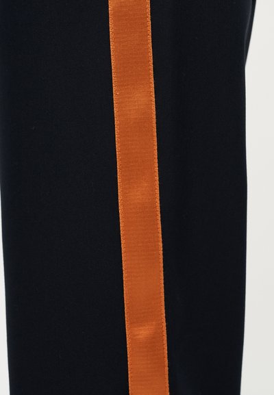 Σκούρο μπλε παντελόνι φόρμας με κάθετη πορτοκαλί ρίγα. Το ύφασμα φαίνεται λείο και ελαστικό, με καθαρό φινίρισμα στις άκρες της ρίγας.