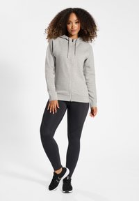 Felpa grigia con zip, cappuccio, maniche lunghe e polsini a costine; abbinata a leggins neri e scarpe da ginnastica nere. Design semplice e aderente.