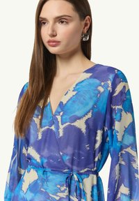 Sukienka z długim rękawem, zapinana w niebiesko-kremowy abstrakcyjny wzór, z głębokim dekoltem w serek i paskiem wiązanym w talii. Gładka tekstura materiału.