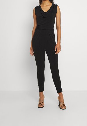 Kobieta ubrana w czarny kombinezon bez rękawów z drapowanym dekoltem i zwężanymi nogawkami, do tego czarne sandały na obcasie z paskami.