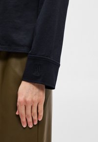 Črna dolgorasna majica z rebrastim ovratnikom in vezenim logotipom. Tkanina se zdi mehka in ima gladko teksturo. Roka počiva na olivno zeleni obleki.