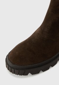 Brun ruskind støvle med rund tå, struktureret skaft og en tyk sort gummisål med et robust mønster.