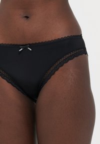 Culotte noire pour femmes, en tissu lisse avec un bord en dentelle. Présente de petits nœuds et le logo HUGO sur la ceinture avant.