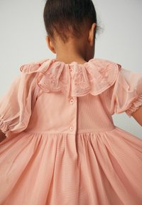 Robe rose avec un col à volants orné de broderies, des manches bouffantes en voile transparent, et une fermeture à boutons au dos. Tissu doux et texturé.
