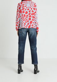 Pitkähihaisen paidan taustana on vaalea sininen, jossa on punaisia ja vaaleanpunaisia epäsäännöllisiä muotoja, yhdistettynä tummansinisiin suoriin farkkuihin ja mustiin nilkkasaappaisiin.