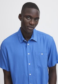 Camisa de botones de manga corta en azul vibrante, hecha de una tela suave, con un bolsillo en el pecho y botones blancos.