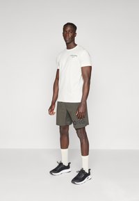 Tricou alb cu mânecă scurtă și text negru, pantaloni șort verde măsliniu cu imprimeu "BORG" și pantofi sport negri cu tălpi albe și șosete deschise.