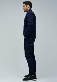 Mann in navy-blauem Pufferjacke, dunklen Jeans und schwarzen Sneakers steht seitlich mit Händen in den Taschen der Jacke vor einem schlichten Hintergrund.