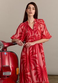 Robe rouge transparente avec motif de feuilles de palmier, manches courtes et larges, et décolleté en V. La mannequin est adossée à un scooter rouge.
