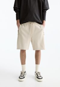 Person wearing beige drawstring shorts, cream socks, black and white sneakers, and a loose black long-sleeve top, standing with hands in pockets.