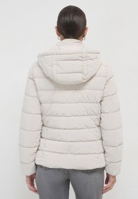 Frau in einer hellbeigen Daunenjacke mit Kapuze und grauen Jeans, von hinten vor einem schlichten weißen Hintergrund gezeigt.