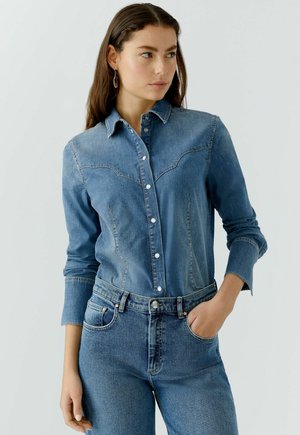 Denim shirt met een knoopsluiting aan de voorkant, puntige kraag en twee borstzakken met gebogen naden. Gekleed met bijpassende blauwe jeans.