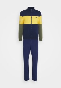 Survêtement homme deux pièces avec veste zippée en bleu marine, jaune et vert olive, assorti à un pantalon bleu marine.