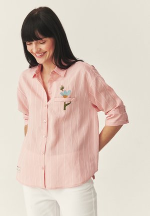 Kobieta uśmiechająca się, ubrana w jasnoróżową koszulę w paski z podwiniętymi rękawami, z małym haftowanym niebieskim kwiatem i pszczołą przy kieszeni na piersi.