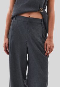 Midja av en person som bär mörkgrå skräddarsydda byxor och en matchande ärmlös topp med spänndetalj, mot en enfärgad ljus bakgrund.