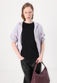 Lichtpaarse ritsjack met opgestikte mouwen over een zwart t-shirt, gecombineerd met zwarte broek en een bordeauxlederen handtas.
