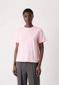 Pink cotton t-shirt with a crew neck and short sleeves, featuring a small white logo on the left chest. Model wears gray high-waisted trousers.