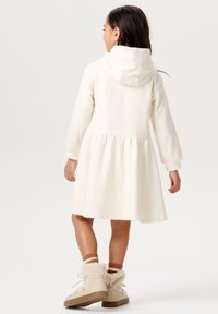 Abito crema con cappuccio, maniche lunghe e vita arricciata. Indossato con stivali beige e calze crema texture. Tessuto morbido, design casual.