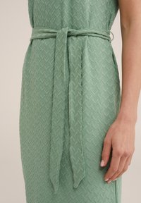 Vihreä kuvioitu mekko, jossa on teksturoitu sahalaita-kuvio. Siihen sisältyy vastaava kangashihna, joka on solmittu vyötärölle, korostaen siluettia.