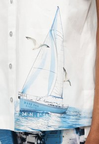 Ιστιοφόρο με το όνομα "Marc Cain Regatta" με μπλε και άσπρα πανιά στο νερό, συνοδευόμενο από δύο πετούμενους γλάρους σε λευκό φόντο.