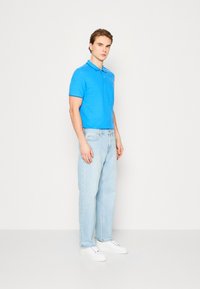 Polo de culoare albastră cu guler și mâneci scurte, asociat cu blugi drepti de culoare albastră deschisă și adidași albi. Elemente de design minime.