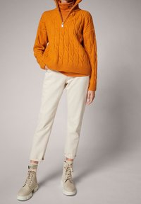Maglione arancione a coste con colletto alto, pantaloni beige chiaro e stivali beige con lacci e suola spessa, posati su uno sfondo neutro.