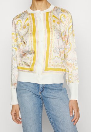 Osoba nosząca zapinany, długi sweter typu kardigan z żółtymi, różowymi i szarymi wzorami kwiatowymi i abstrakcyjnymi, w połączeniu z jasnoniebieskimi dżinsami.