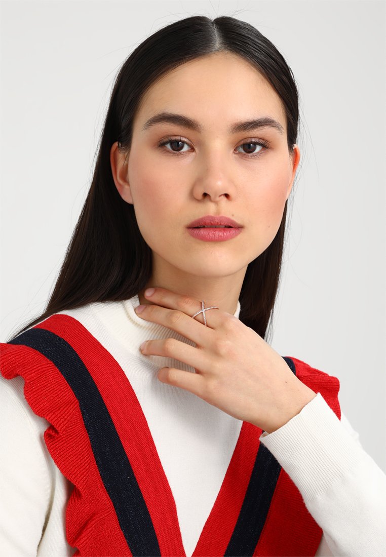 Witte trui met rode geplooide accenten en donkere strepen, model draagt een zilveren kruisring, glad haar, neutrale make-up, gecentreerd.