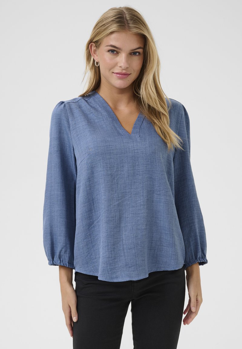 Blusa azzurra, leggera, con maniche lunghe e scollo a V, finitura texture e polsini arricciati, abbinata a pantaloni neri.