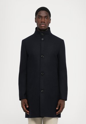 Cappotto di lana blu navy con colletto alto, caratterizzato da sei bottoni neri e due tasche laterali, vestibilità sartoriale e texture liscia.
