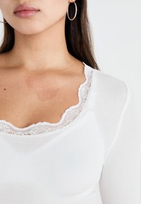Valge ribaga pikkade varrukatega topp, millel on pitsiga ääristatud kaelus, näidates pehmet tekstuuri ja vormivat siluetti.
