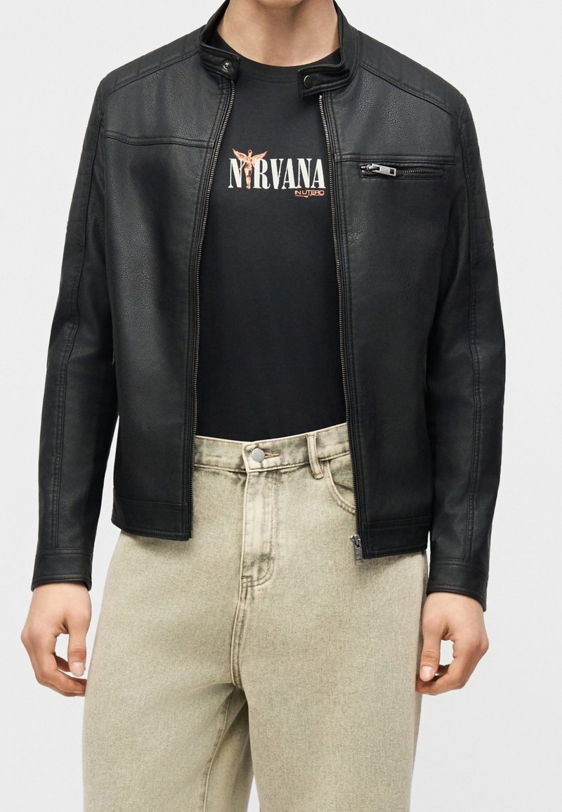 Personne portant une veste en cuir noire ouverte sur un t-shirt noir avec un motif Nirvana et un jean beige clair taille haute, coupé au niveau du cou et à mi-cuisse.