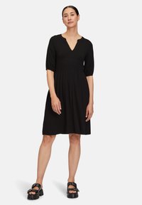 Schwarzes Kleid mit kurzen Puffärmeln, gerafftem Taillenbund und V-Ausschnitt. Getragen mit klobigen schwarzen Sandalen mit Schnallenakzenten.