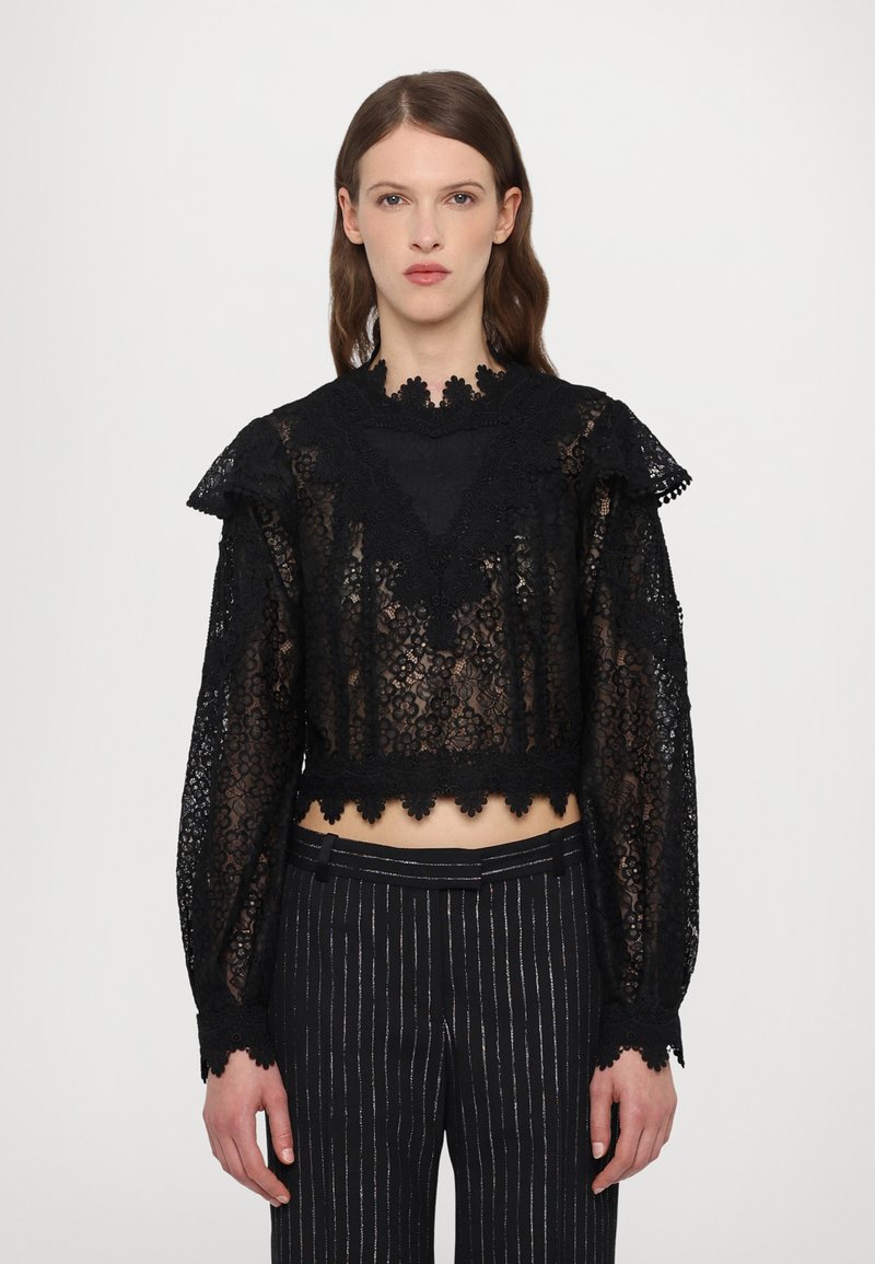 Black lace cropped top with floral patterns, ruffled shoulders, and a scalloped hem. Long sleeves with intricate detailing. Paired with striped pants.