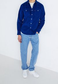 Blå kordtøjskjorta med knapper och två bröstfickor, kombinerad med ljusblå jeans och vita sneakers. Modellen står mot en vit bakgrund.