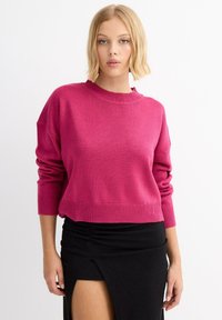 Mujer con cabello rubio corto que lleva un suéter rosa brillante y una falda negra con una abertura frontal, sobre un fondo blanco.