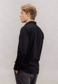 Svart langermet skjorte laget av glatt stoff, med klassisk krage, knappeermer og en skreddersydd passform, vist fra siden.