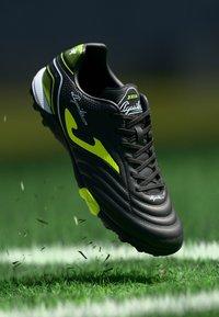 Μαύρο ποδοσφαιρικό παπούτσι με πράσινες και λευκές λεπτομέρειες, επάνω μέρος από υφή δέρματος, σχέδιο με κορδόνια και κίτρινη σόλα. Πάνω από γκαζόν.