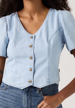 Femme portant un haut en jean bleu clair à manches courtes boutonné avec des épaules bouffantes et un jean bleu foncé taille haute.