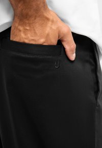 Černé látkové kalhoty s hladkou texturou, s boční kapsou a minimalistickým brandingem. Ruka zasunutá v kapse.