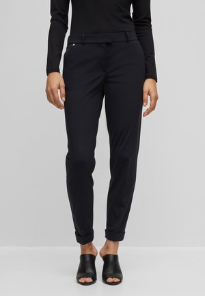 Femme portant un pantalon noir ajusté avec des revers et des mules à talons noires ouvertes, debout devant un fond gris clair uni.
