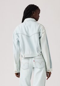 Chaqueta de mezclilla azul claro con un diseño clásico, que presenta un canesú puntiagudo, cierre de botones y costuras en contraste. Textura suave y forma ajustada.