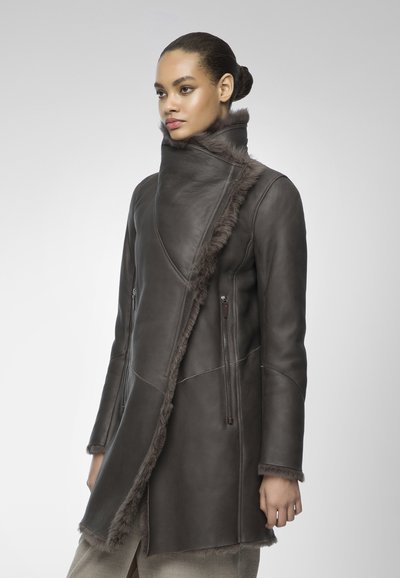 Cappotto di pelle marrone scuro con colletto alto, con rifiniture in pelliccia e tasche laterali con zip. Texture liscia con vestibilità su misura e dettagli delle cuciture angolari.